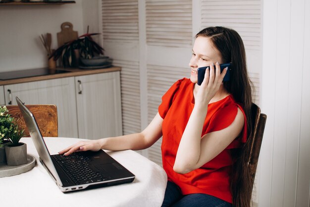 chica con una blusa roja está sentada en una computadora portátil en casa y hablando por teléfono