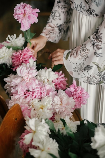 Foto chica bohemia sosteniendo peonías rosadas y blancas en las manos en una silla de madera rústica mujer hipster con estilo en vestido bohemio arreglando flores de peonía decoración de boda del día internacional de la mujerxa