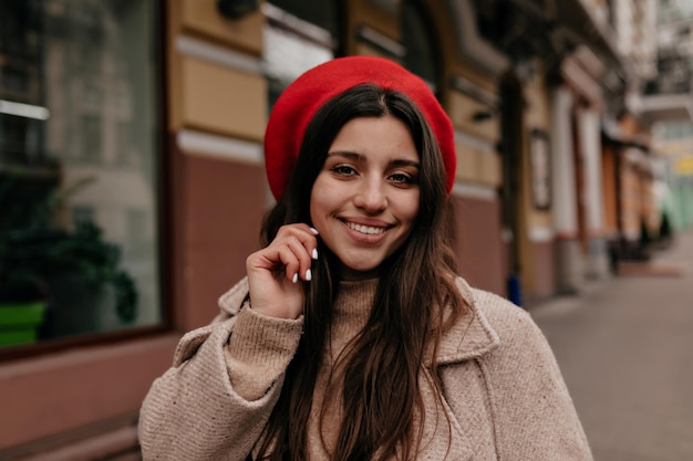 Una chica bonita y alegre con una gran sonrisa cerrada, cabello largo y oscuro con una elegante boina roja, sonríe y posa contra el fondo del centro de la ciudad