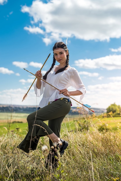 Chica bonita cazadora disparando arco y flecha en la pradera