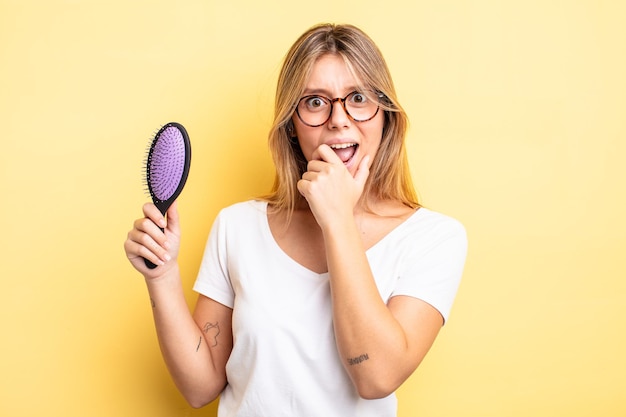 Chica bonita rubia con la boca y los ojos bien abiertos y la mano en la barbilla. concepto de cepillo de pelo