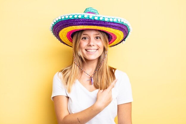 chica bonita rubia sonriendo alegremente, sintiéndose feliz y apuntando hacia un lado. concepto de sombrero mexicano