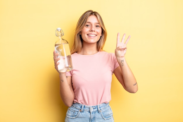 Chica bonita rubia sonriendo y mirando amigable, mostrando el número tres. concepto de botella de agua