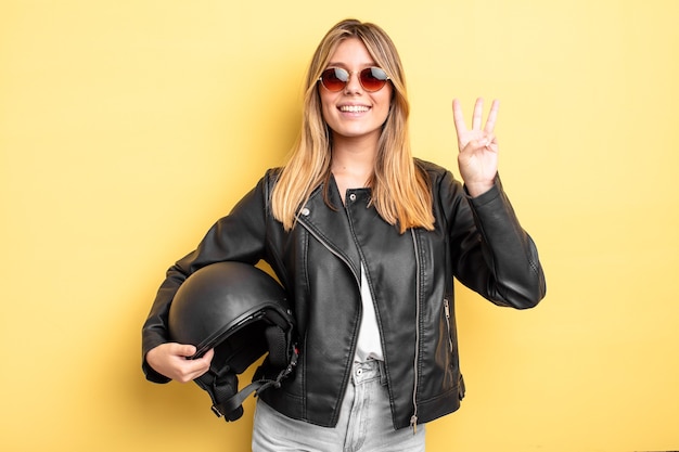 Chica bonita rubia sonriendo y mirando amigable, mostrando el número tres. concepto de casco de moto