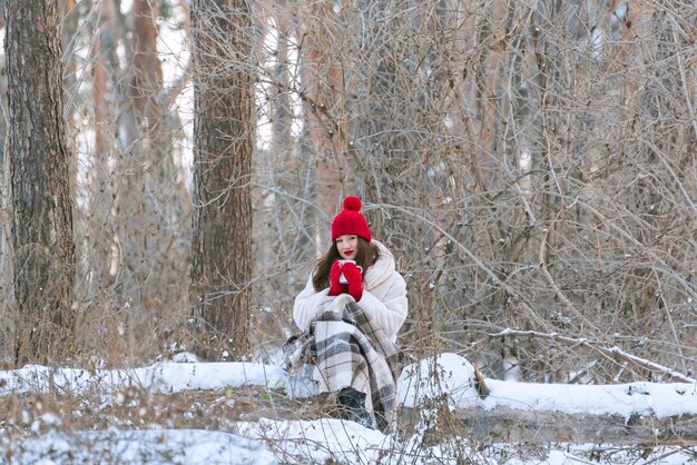 Chica en el bosque de invierno bebe té envuelto en una manta. Mujer con sombrero rojo en manada nevada. copia espacio