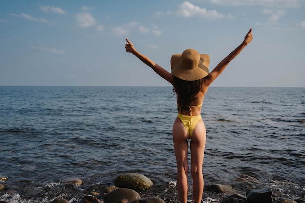 Chica con un botín sexy con un sombrero de paja en traje de baño levanta las manos mientras está de espaldas en la playa junto al mar