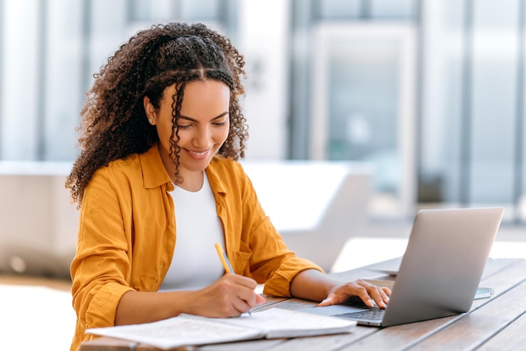 Foto chica brasileña o latina inteligente, exitosa, mujer independiente o estudiante vestida con una camisa naranja, usa una computadora portátil para trabajar o estudiar en línea mientras está sentada al aire libre, toma notas en una sonrisa de cuaderno