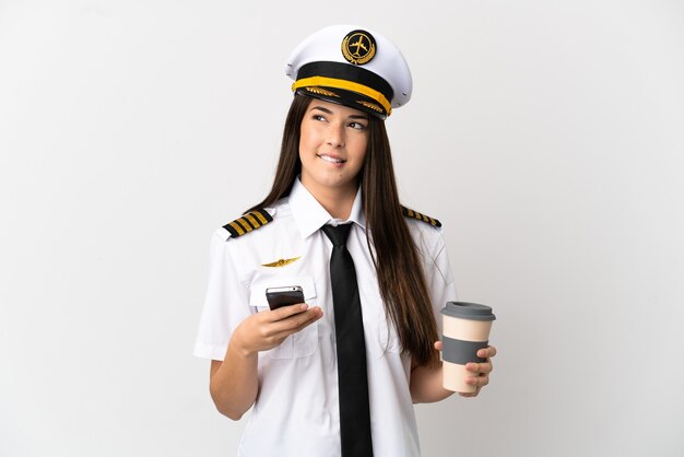 Chica brasileña piloto de avión sobre fondo blanco aislado sosteniendo un café para llevar y un móvil mientras piensa en algo