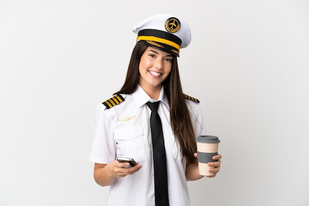 Chica brasileña piloto de avión sobre fondo blanco aislado sosteniendo café para llevar y un móvil