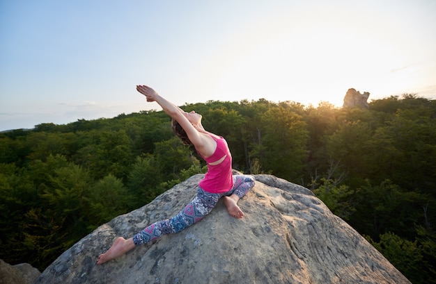 Chica con los brazos levantados haciendo ejercicios de yoga estiramiento sobre roca enorme