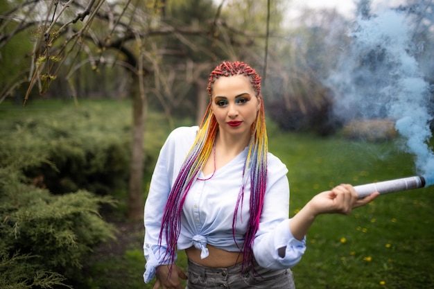 Una chica brillante con maquillaje brillante y trenzas africanas con una camisa azulada se refugia en el humo artificial en un parque lleno de árboles verdes.