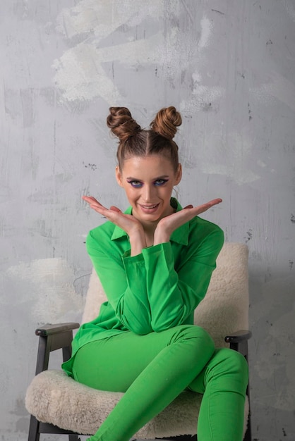 Chica brillante con maquillaje de noche en traje de pantalón verde se sienta en una silla sobre fondo de pared gris Marco vertical