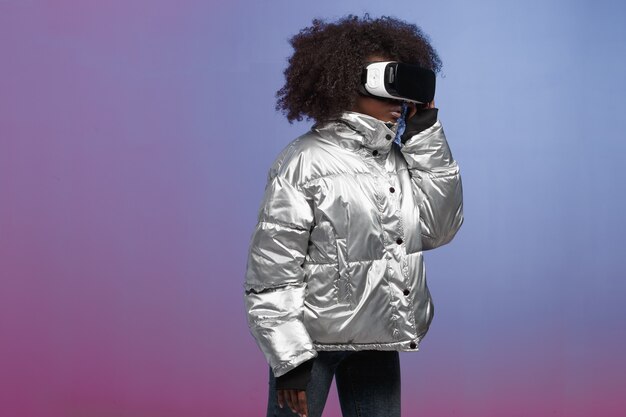 La chica de cabello castaño rizado de moda vestida con una chaqueta de color plateado usa las gafas de realidad virtual en el estudio sobre fondo de neón.