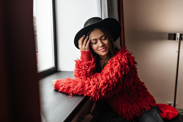 Chica con cabello lacio oscuro toca su rostro Retrato de mujer con sombrero negro y chaqueta roja posando cerca de la ventana