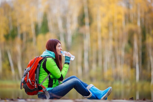 Chica con cabello largo bebiendo agua con mochila sentado entre árboles de otoño