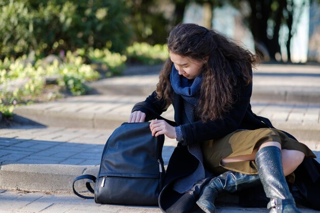 Una chica con cabello largo está sentada en los escalones y tratando de encontrar las cosas correctas en su bolso.