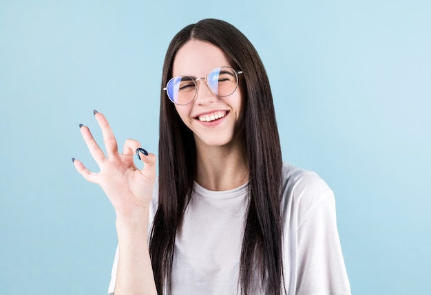 Chica con cabello largo y negro guiñando un ojo, sonriendo y mostrando el signo de OK sobre backgrund azul.