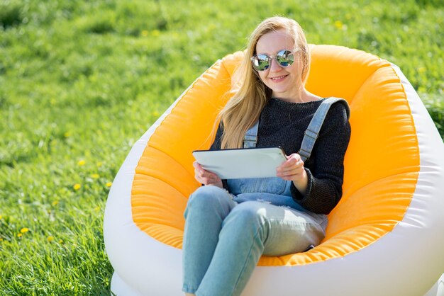 Chica con cabello largo rubio con una tableta sentada en una silla inflable en un césped verde