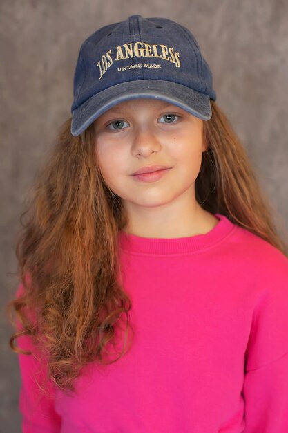 una chica de cabello marrón con cabello rizado en un traje de entrenamiento rosa y una gorra de béisbol azul en la cabeza