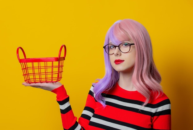 Chica con cabello morado y suéter rojo mantenga la cesta de la compra sobre fondo amarillo