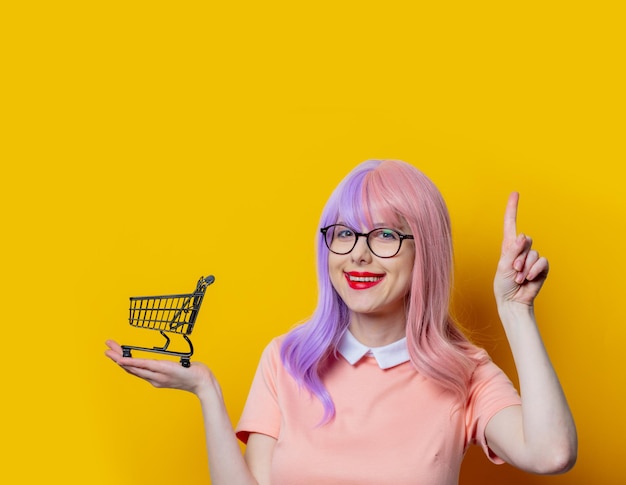 Chica con cabello morado y vestido rosa con carrito de compras negro sobre fondo amarillo