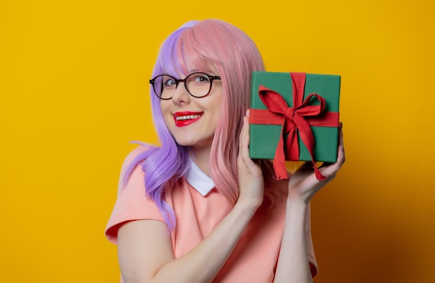 Chica con cabello morado y vestido rosa mantenga caja de regalo sobre fondo amarillo