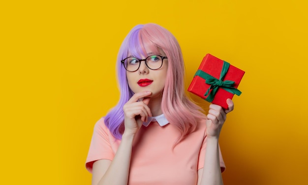 Chica con cabello morado y vestido rosa mantenga caja de regalo sobre fondo amarillo