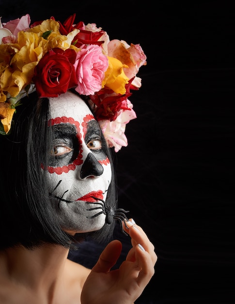 Chica con cabello negro está vestida con una corona de rosas multicolores y maquillaje en su rostro calavera de azúcar