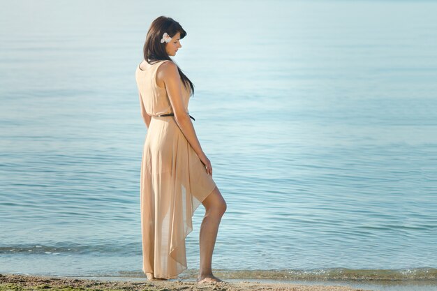 Chica con cabello negro con un vestido beige en la orilla del mar azul