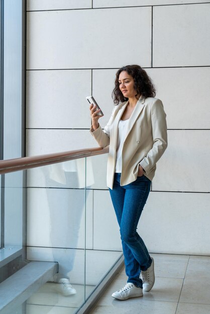 Una chica de cabello oscuro con una chaqueta ligera y jeans azules se para cerca de una ventana grande con un teléfono inteligente Mira el teléfono