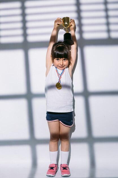 Foto chica de cabello oscuro con medallas de campeonato deportivo, levantando su copa con las manos, por encima de las sombras de una ventana con persiana encima de ella. concepto de deporte y victoria