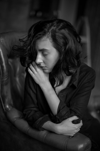 Chica con cabello oscuro rizado y maquillaje natural sentada en una silla de cuero oscuro foto en blanco y negro