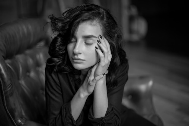 Chica con cabello oscuro rizado y maquillaje natural sentada en una silla de cuero oscuro foto en blanco y negro