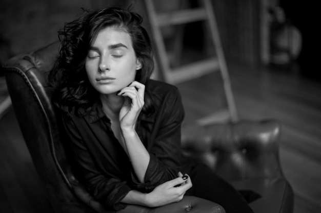 Chica con cabello oscuro rizado y maquillaje natural sentada en una silla de cuero oscuro foto en blanco y negro