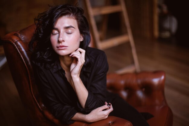 Chica con cabello oscuro rizado y maquillaje natural sentada en una silla de cuero procesamiento de fotos vintage