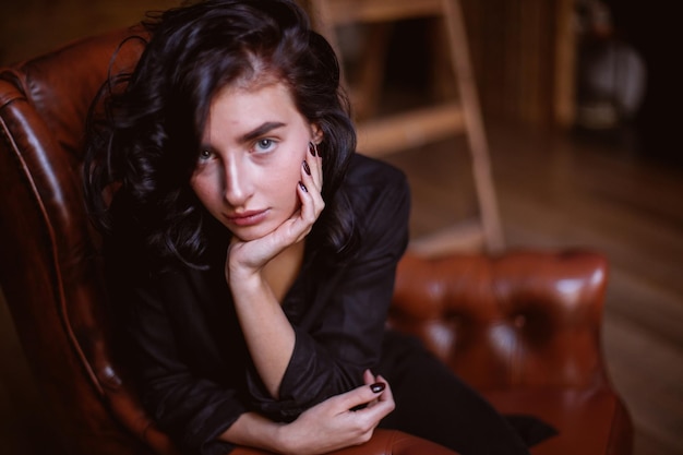 Chica con cabello oscuro rizado y maquillaje natural sentada en una silla de cuero procesamiento de fotos vintage