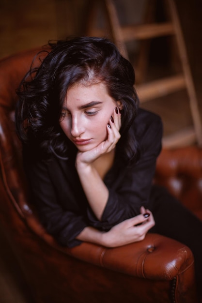 Chica con cabello oscuro rizado y maquillaje natural sentada en una silla de cuero procesamiento de fotos vintage