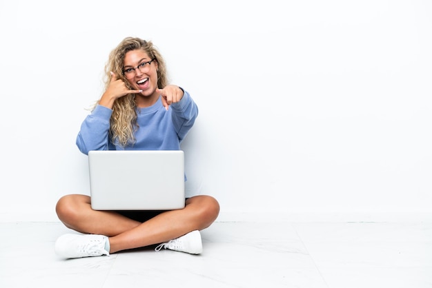 Chica con cabello rizado con una computadora portátil sentada en el suelo haciendo gestos de teléfono y apuntando al frente