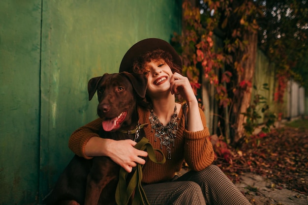 chica con cabello rizado y ropa vintage se sienta en el suelo cerca de una casa de campo abraza al perro