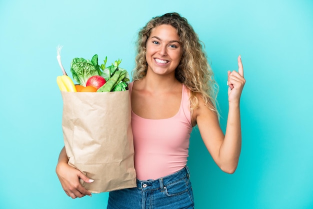 Chica con cabello rizado sosteniendo una bolsa de compras aislada sobre fondo verde apuntando hacia una gran idea