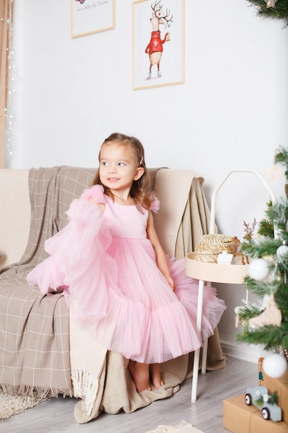 Chica con cabello rubio en un elegante vestido rosa cerca del árbol de Navidad se sienta en el sofá y juega