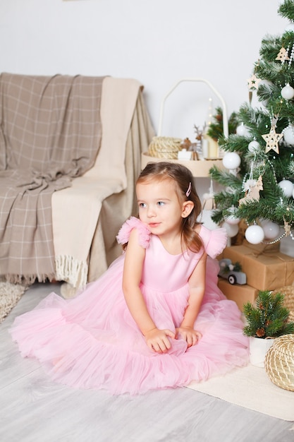 Chica con cabello rubio en un elegante vestido rosa cerca del árbol de Navidad se sienta en el suelo