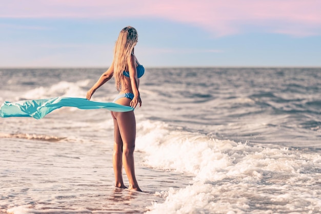 Una chica con cabello rubio en un traje de baño azulado y un mantón brillante camina por la playa