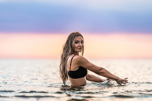 Una chica con cabello rubio en un traje de baño negro salpica a los lados mientras se sienta en un estuario sobre un fondo de puesta de sol