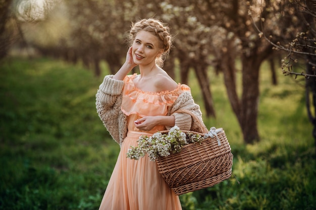 Chica con cabello rubio con un vestido ligero en jardín de flores. Chica en un hermoso vestido y suéter de punto disfruta de la puesta de sol en un jardín de flores de pera, con una canasta de flores