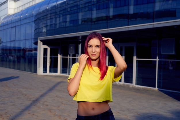 Foto chica con cabello teñido de color rosa y camiseta amarilla se encuentra contra el fondo del edificio de cristal