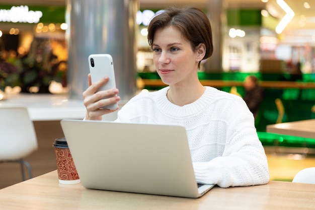 Chica en un café se sienta en una computadora portátil, mira la pantalla del teléfono. Freelancer morena en suéter blanco.