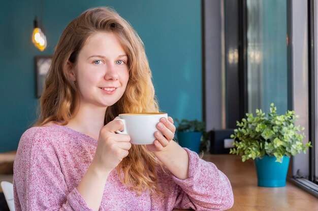 Chica en un café con una taza de café, sonriendo y bebiendo un café con leche.