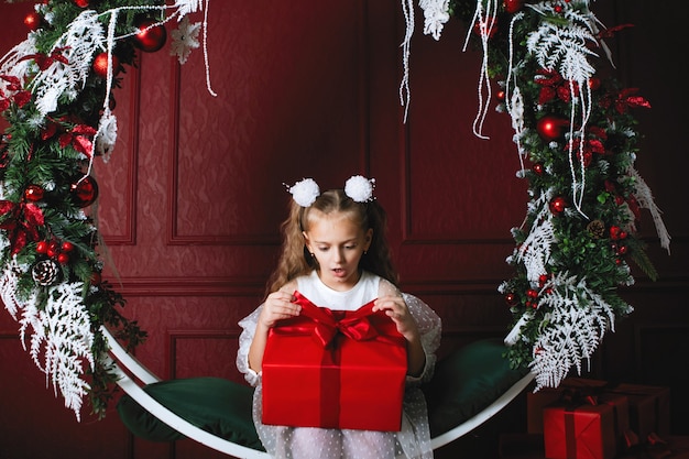 Chica de la caja roja. Regalo de Navidad. Adolescente se sienta en el arco columpio con coronas de decoración de Año Nuevo, ramas de abeto, bolas y guirnaldas.