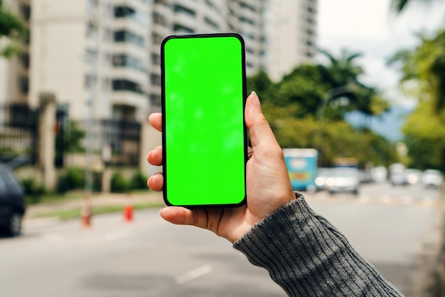 Chica en la calle sosteniendo un teléfono inteligente con pantalla verde esperando que llegue el auto Chroma Key